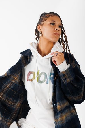 Fashion model in white hoodie and plaid jacket poses against white backdrop in studio setting.