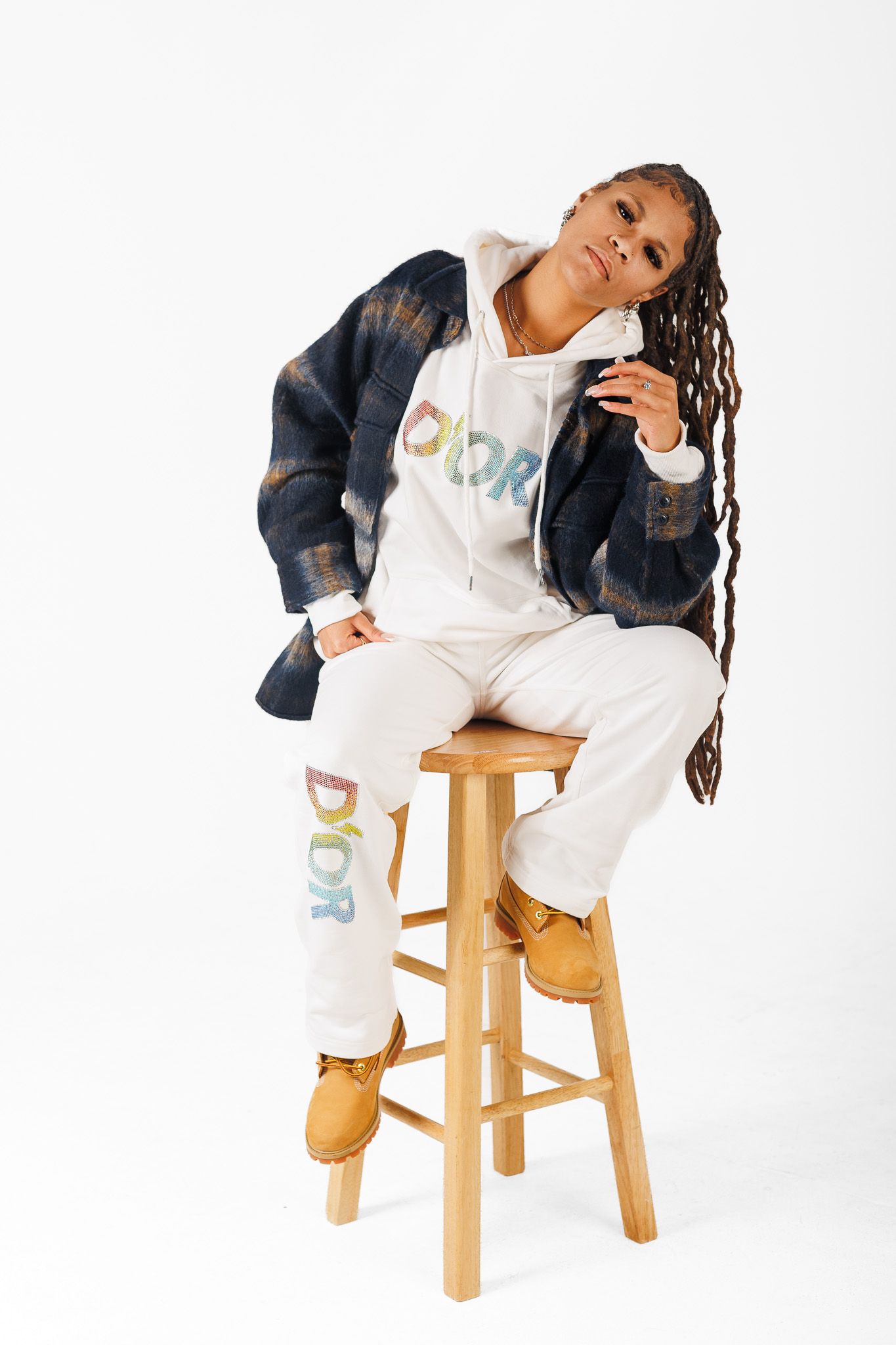 Model in white sweatsuit and denim jacket poses casually on wooden stool against white backdrop.