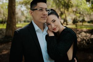 A loving couple in formal attire poses together outdoors in a sunlit park setting during a portrait session.