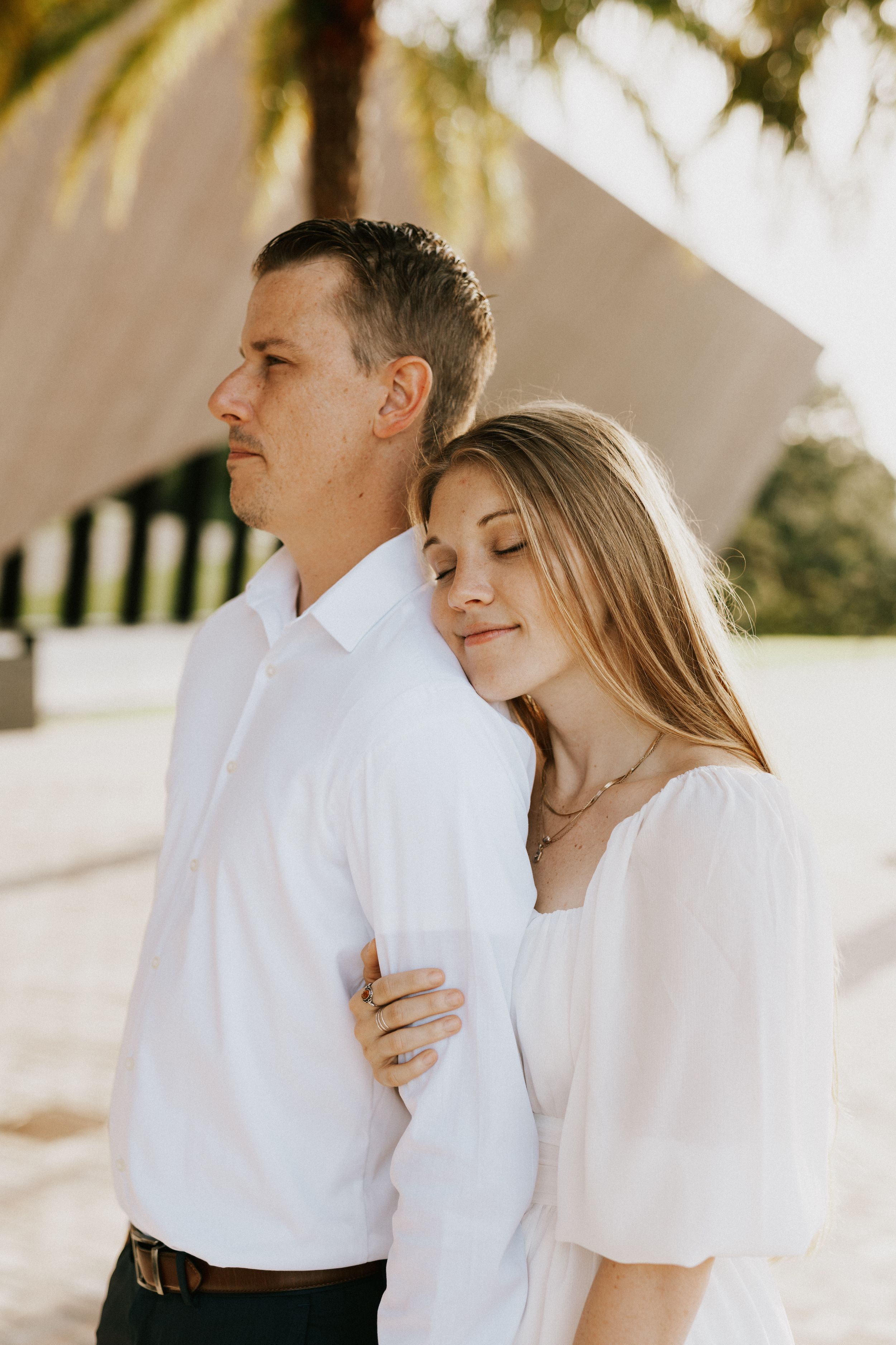 A couple in white outfits embraces tenderly in a sunlit outdoor setting with architectural elements visible.