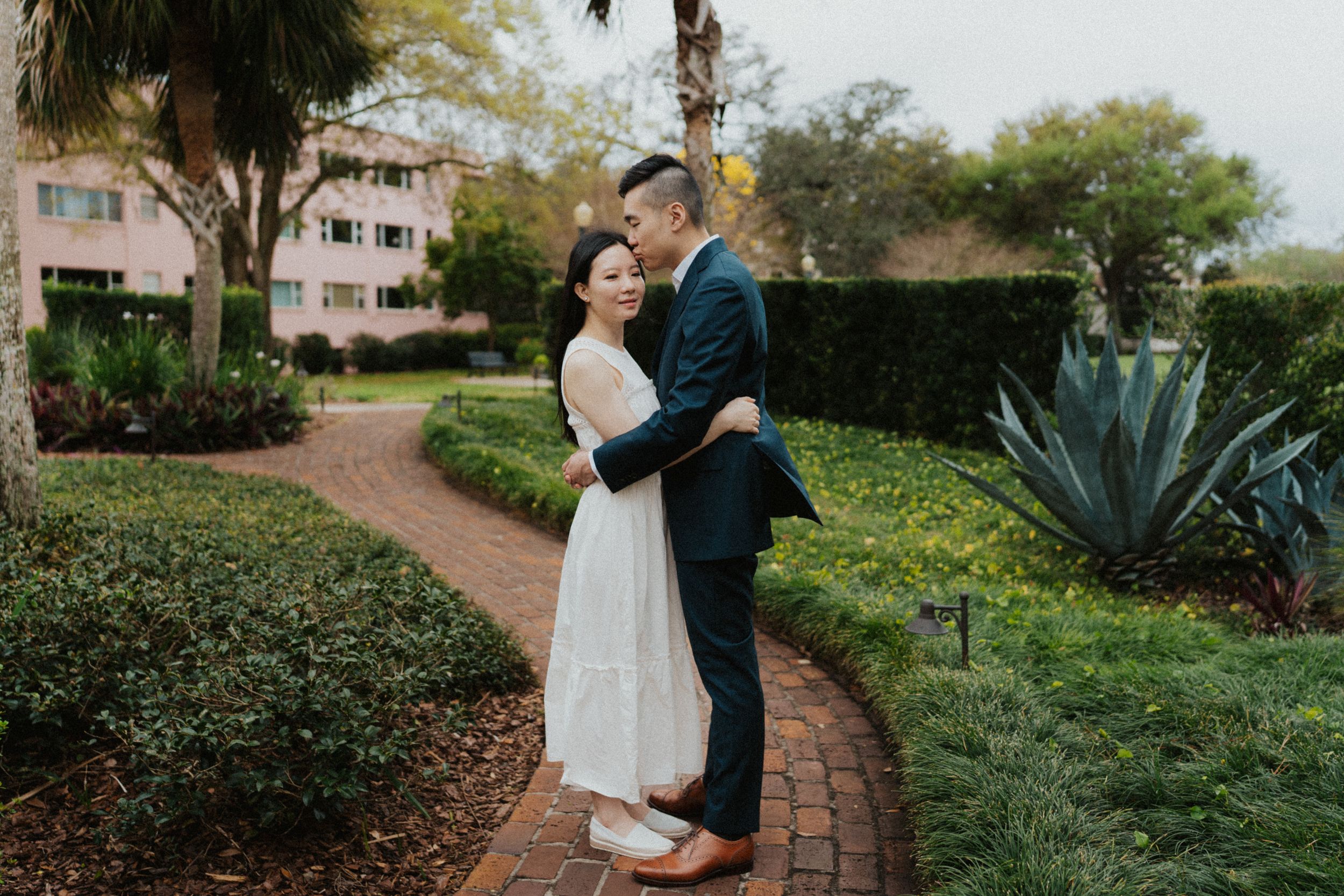 A couple embraces on a curved garden path surrounded by lush greenery and agave plants at an estate property.