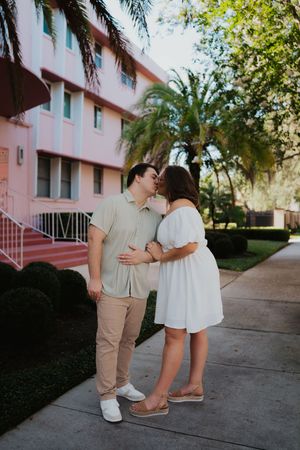 A romantic couple in white and beige outfits sharing a tender moment outside a pink building with palm trees.