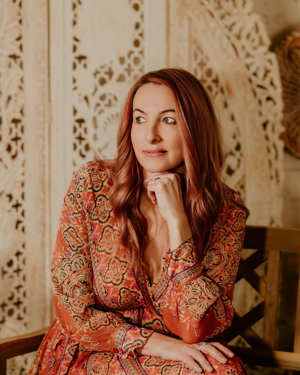 Woman in vintage-inspired orange and brown patterned dress poses against ornate white decorative wall background.