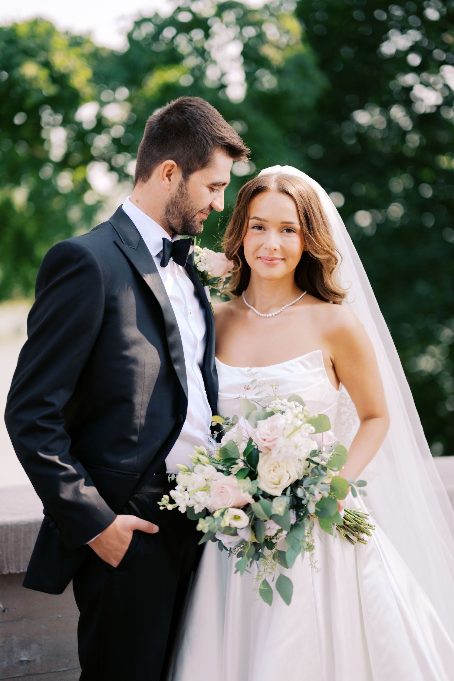 A bride in white gown holds a lush bouquet of white flowers and greenery while standing with groom in black tuxedo outdoors.