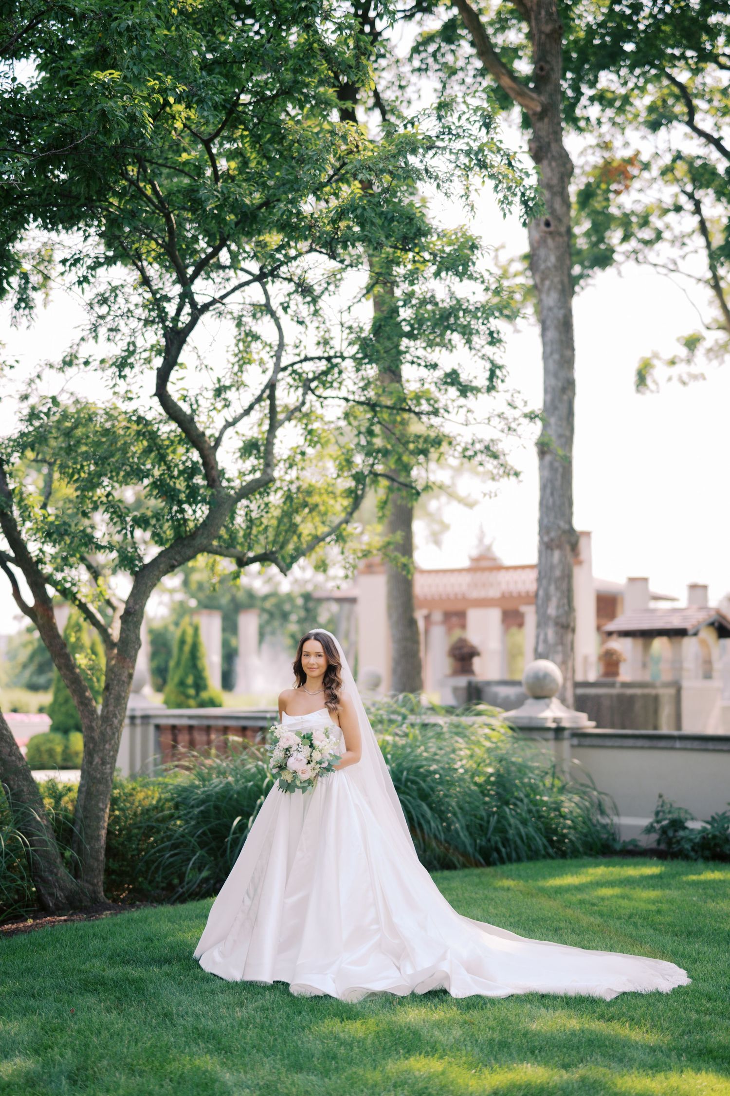 A bride in a flowing white wedding gown poses gracefully in a garden setting with lush trees and greenery.
