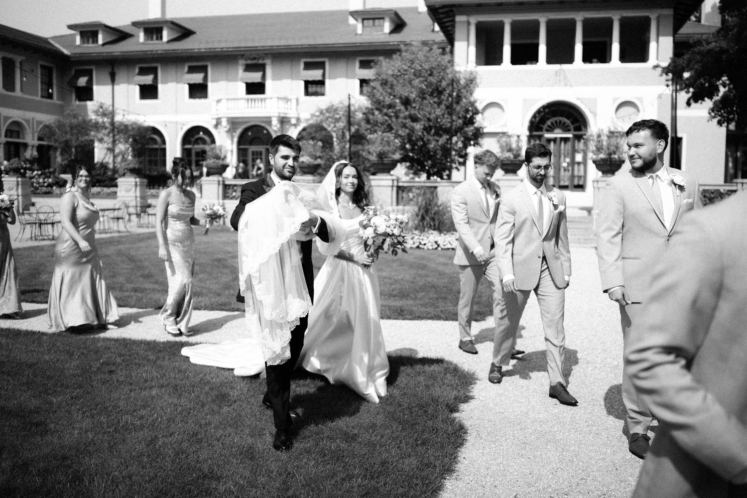 A black and white series of wedding party photos taken outdoors on a lawn near traditional buildings.