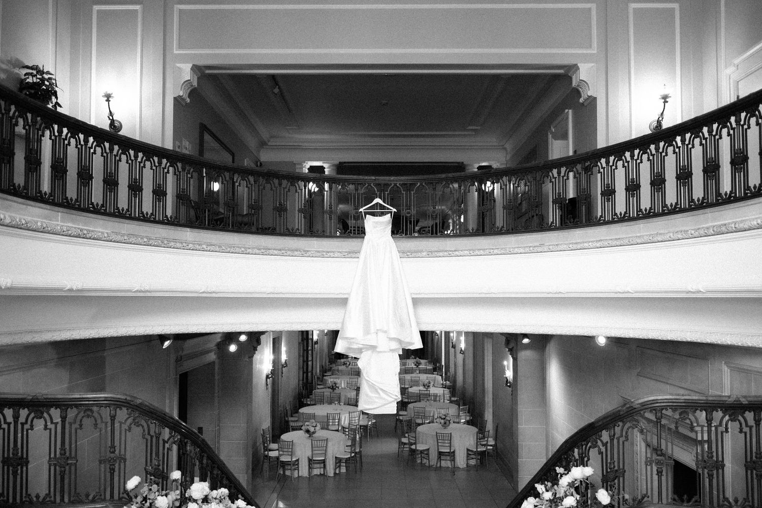 A wedding dress hangs elegantly from a balcony railing in a grand black and white venue with ornate architectural details.