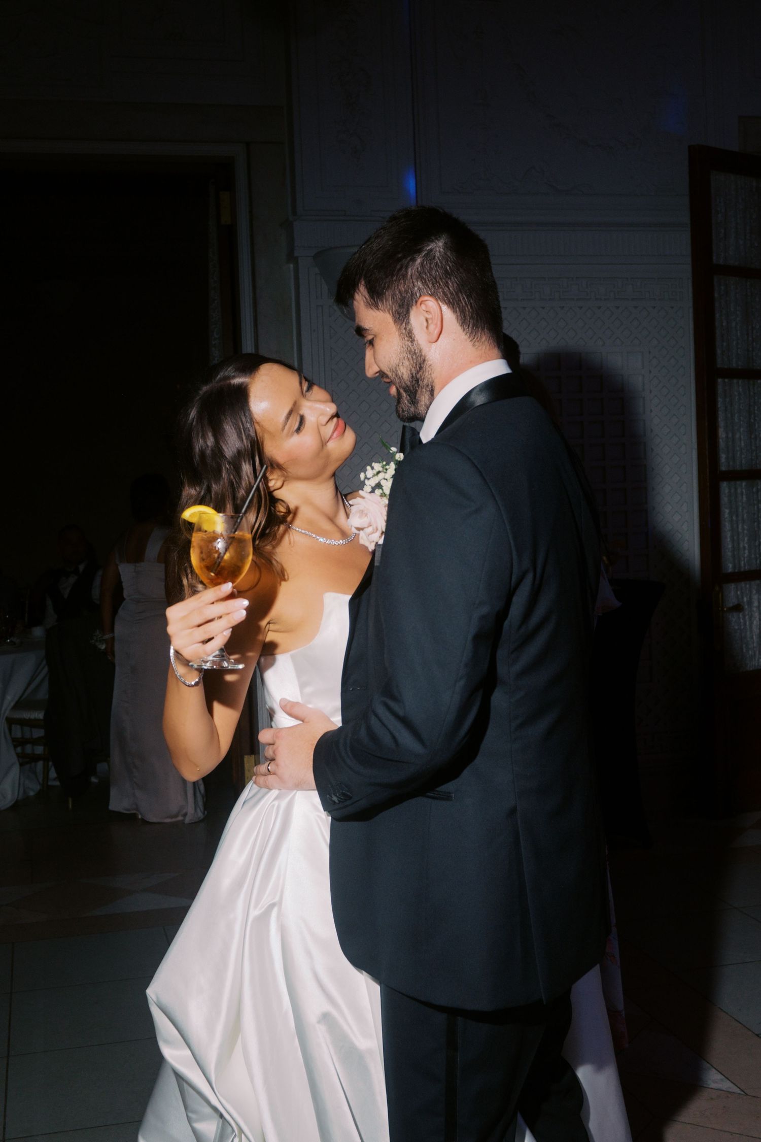 A couple embraces in a dimly lit room during what appears to be a wedding celebration.