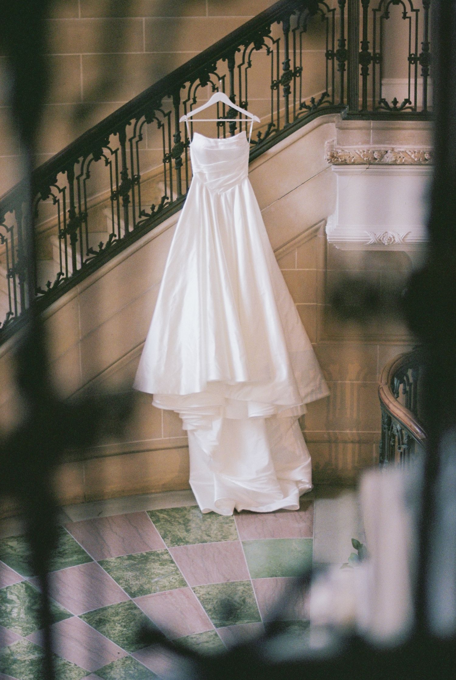 A white wedding dress hangs elegantly against an ornate staircase with wrought iron railings in a classic interior setting.