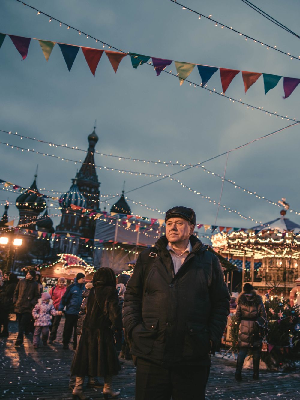 People gather at a festive outdoor winter market with colorful bunting and twinkling lights in Moscow's Red Square at dusk.