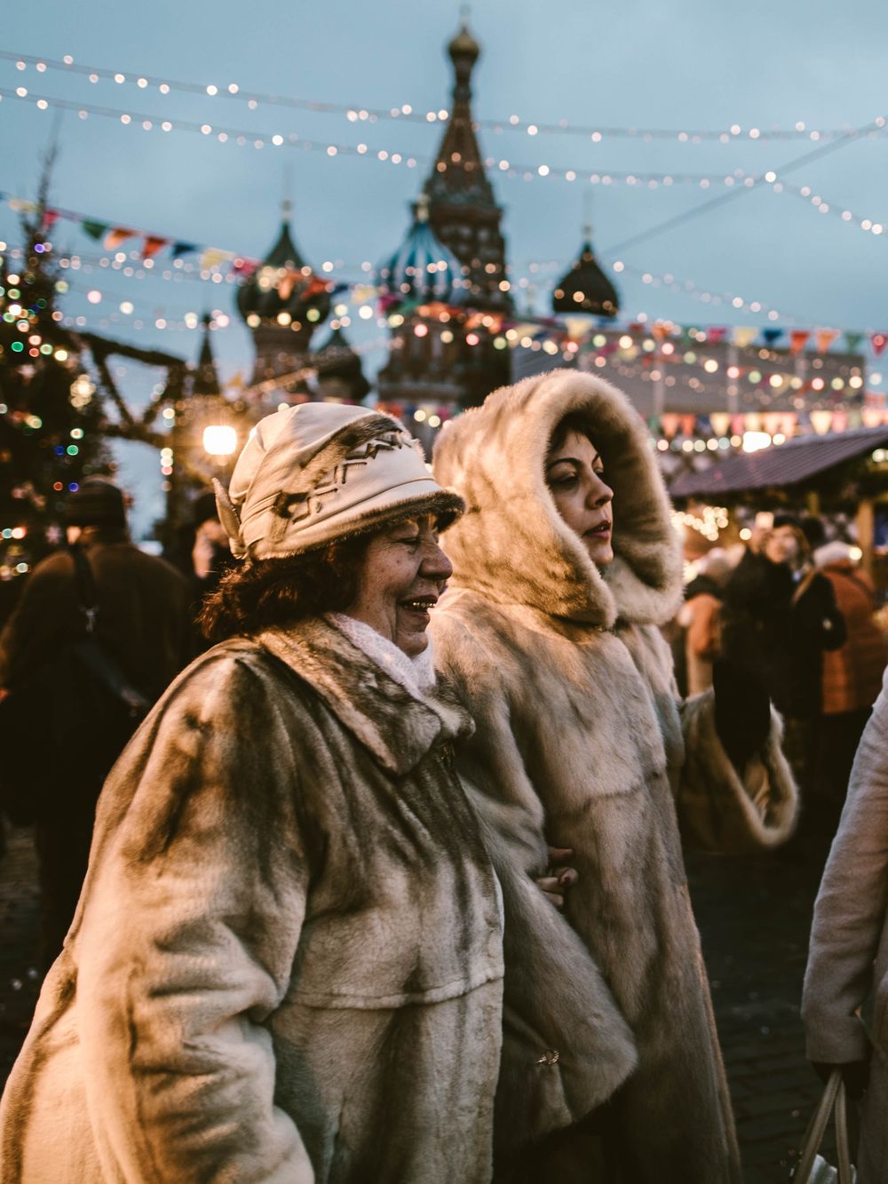 People in winter coats and hats gather at a festive outdoor market illuminated by twinkling string lights at dusk.