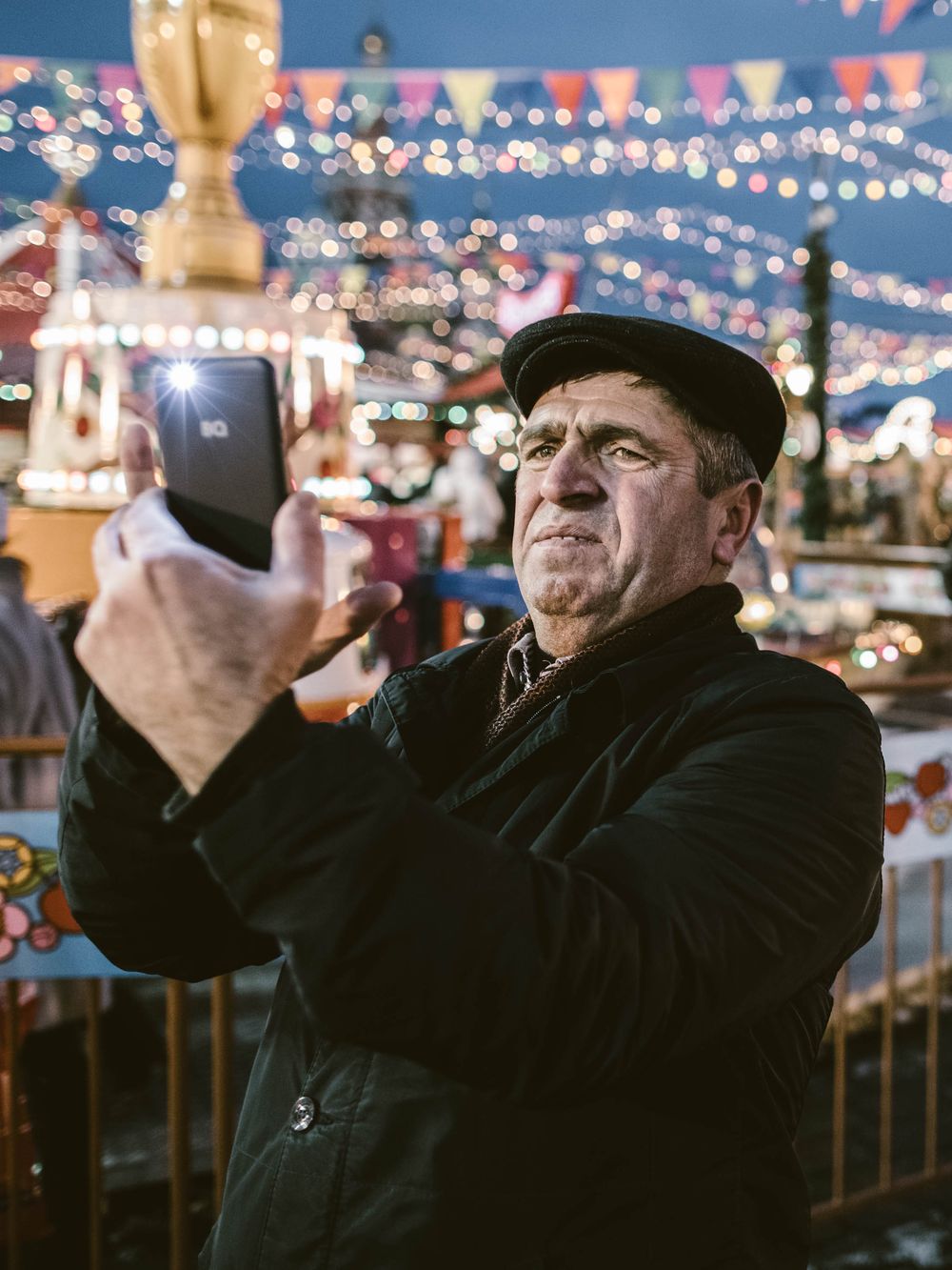 Person in dark coat and hat takes photos with string lights and festive bunting decorations in evening setting.