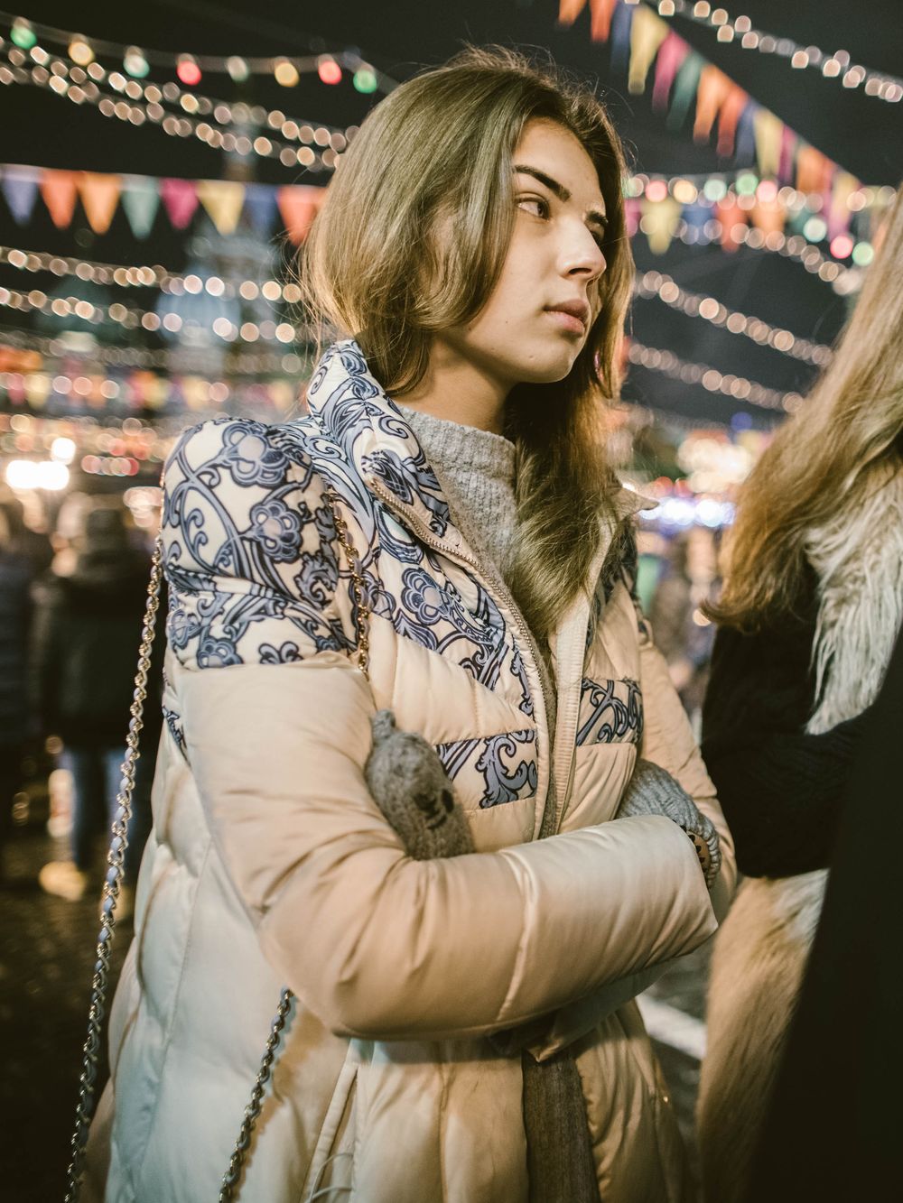 Person wearing a beige winter puffer jacket with floral print stands at night under festive string lights at an outdoor event.