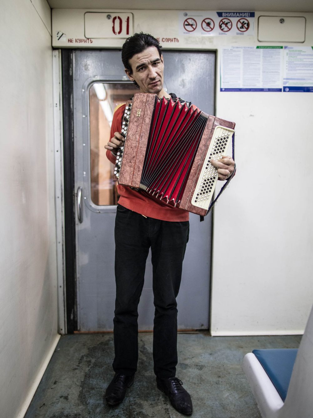 A musician plays a red and white accordion while standing in front of a subway train door.