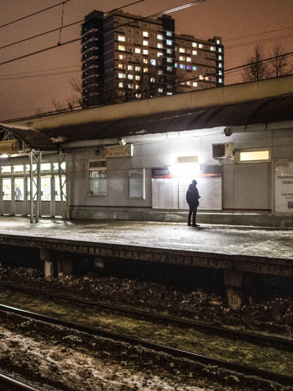 Empty train platform at night with snow on the ground and apartment buildings visible in the distance.