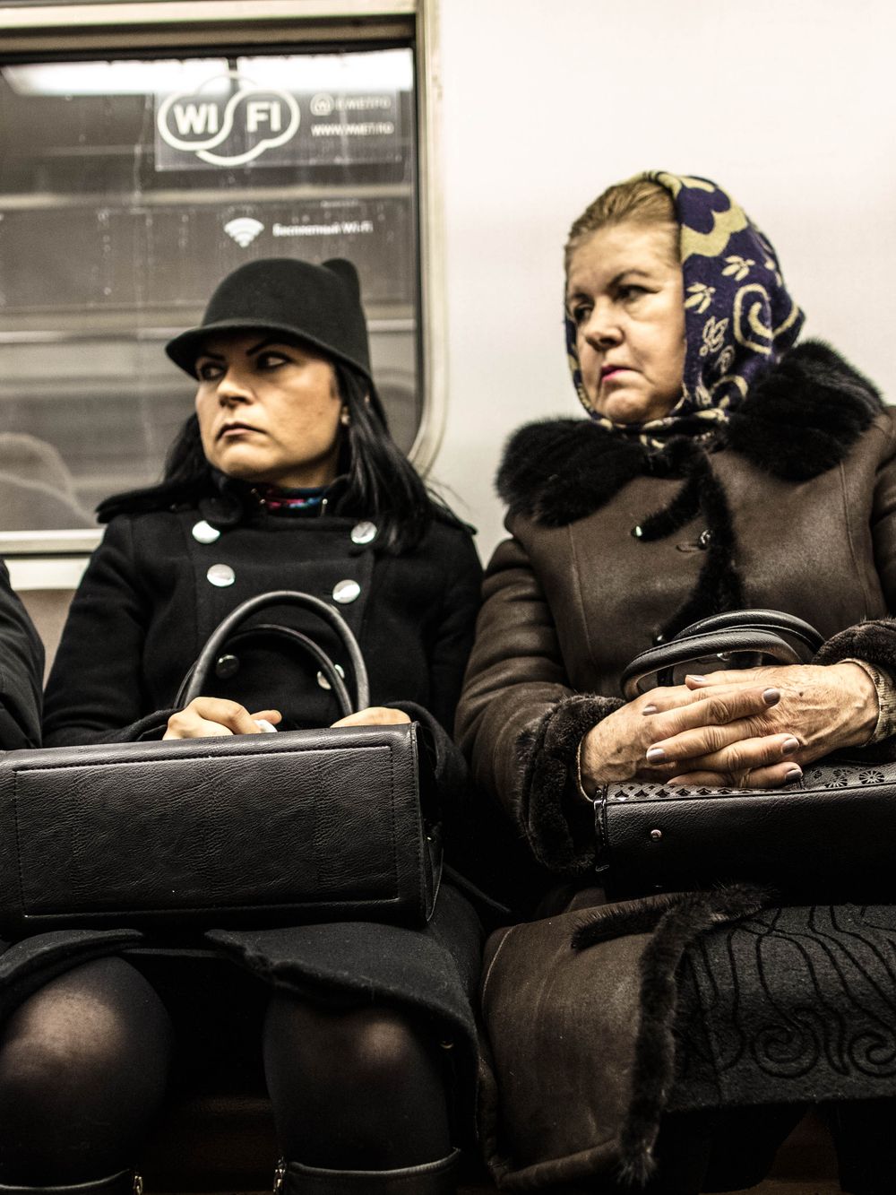 Two passengers sit side by side on public transit, bundled in winter coats and holding bags.