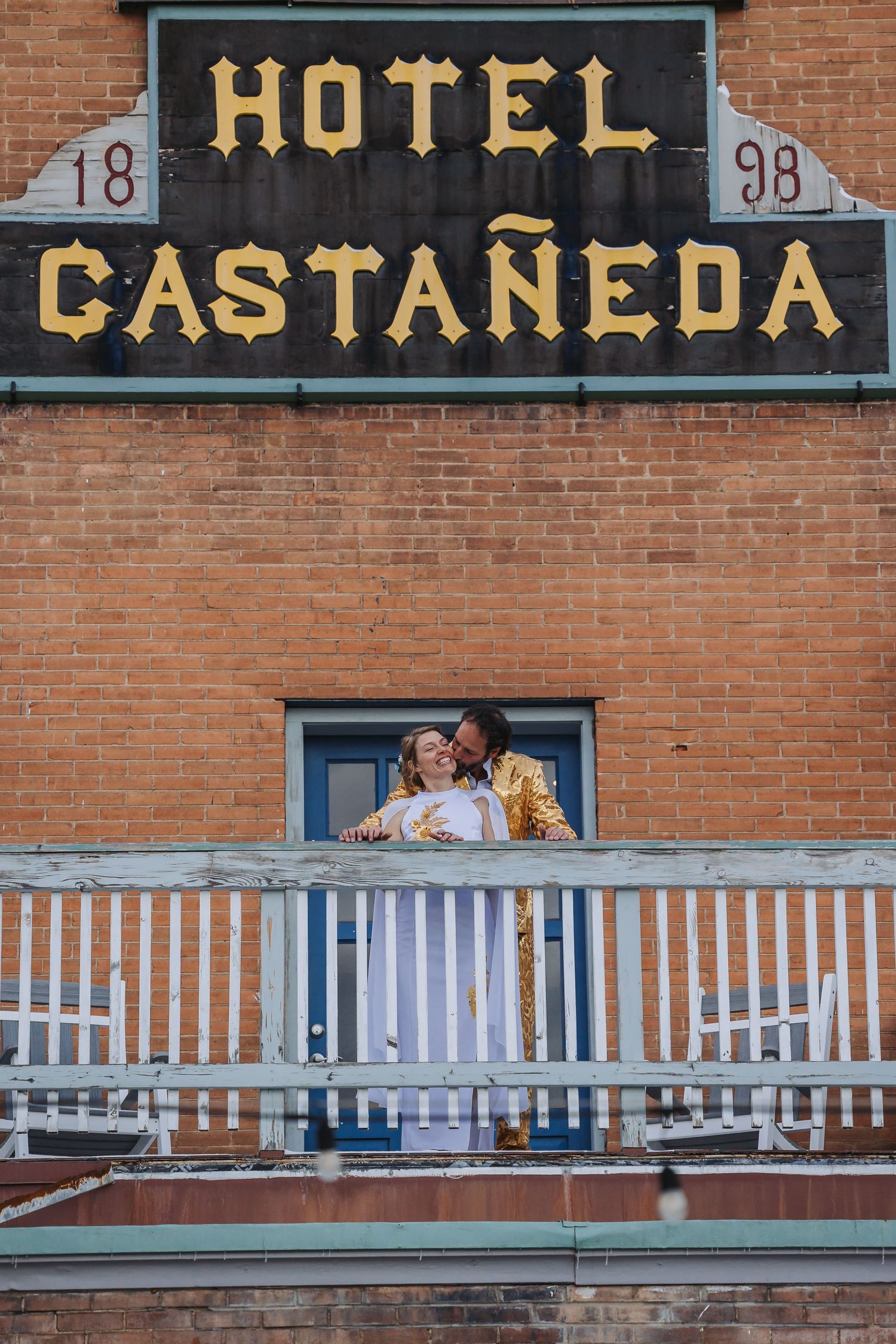 A couple shares an intimate moment on a balcony at the historic Hotel Castaneda with brick walls and white railings.