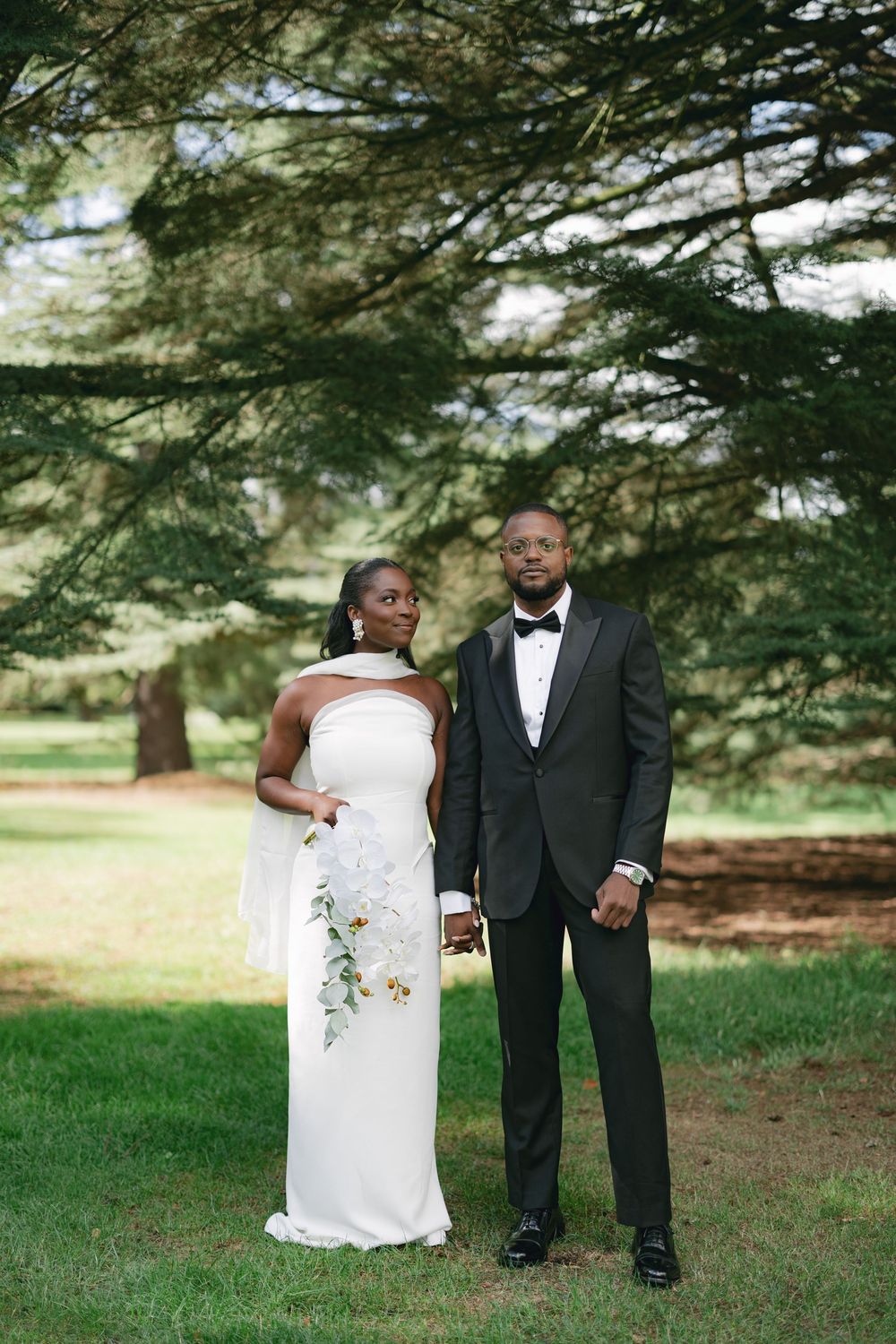 A couple in formal wedding attire poses together on a grassy lawn under evergreen trees.