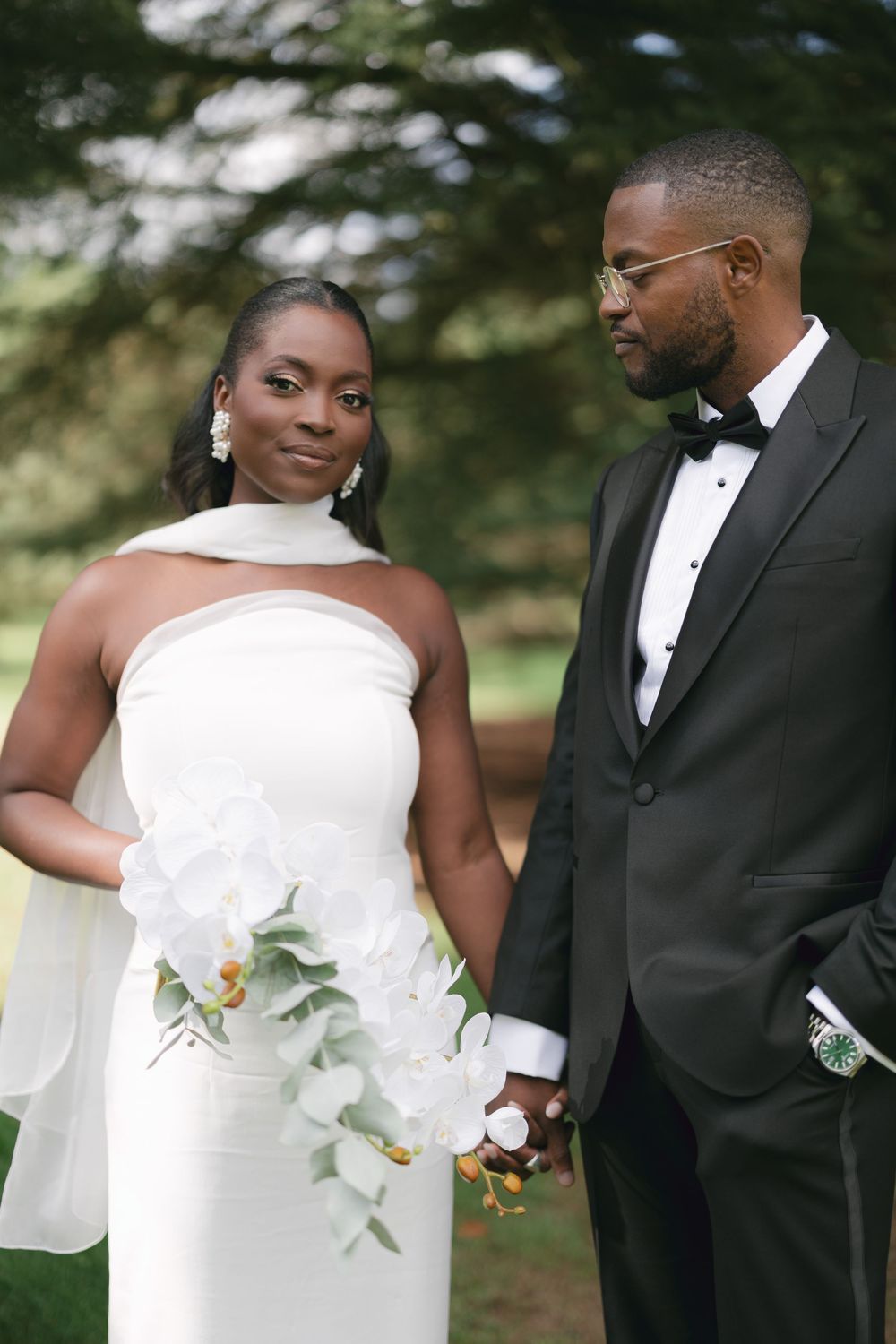 Elegant couple in wedding attire share an intimate moment outdoors with white floral bouquet.