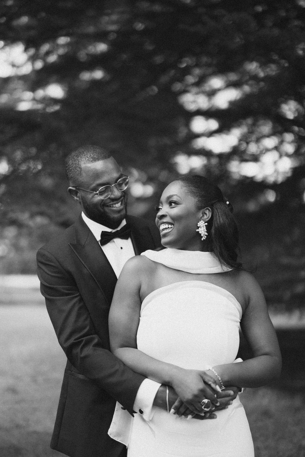 A romantic black and white wedding portrait captured under trees in a garden setting.