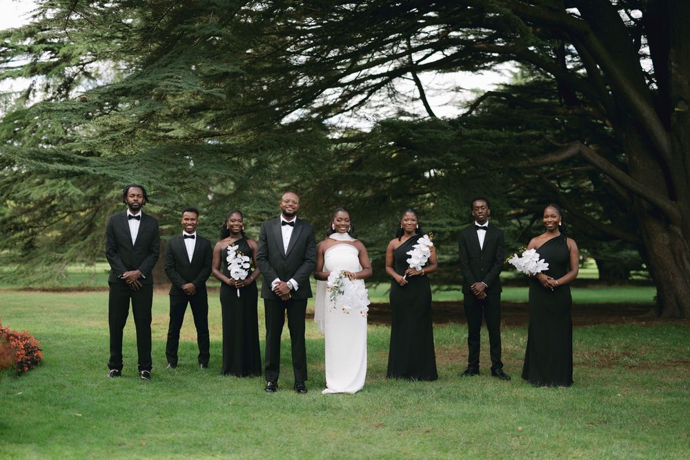 Wedding party in formal attire poses in line against backdrop of tall green trees in outdoor garden setting.