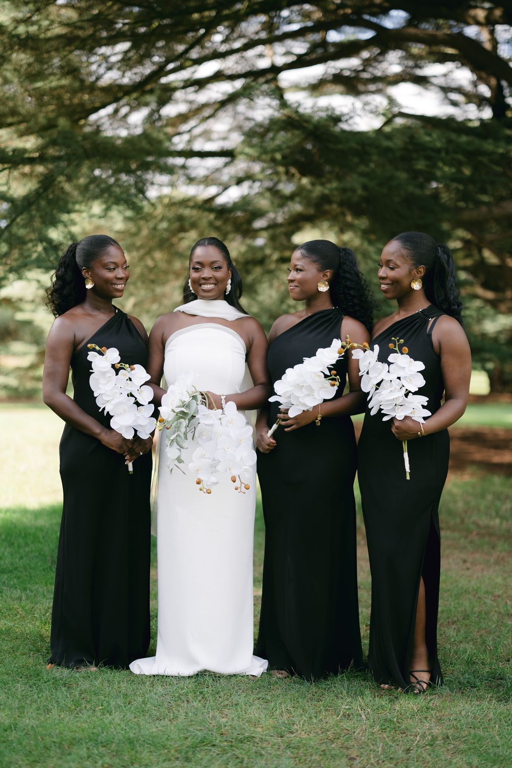 Bridesmaids in black dresses surround bride in white, all holding bouquets under shaded trees.