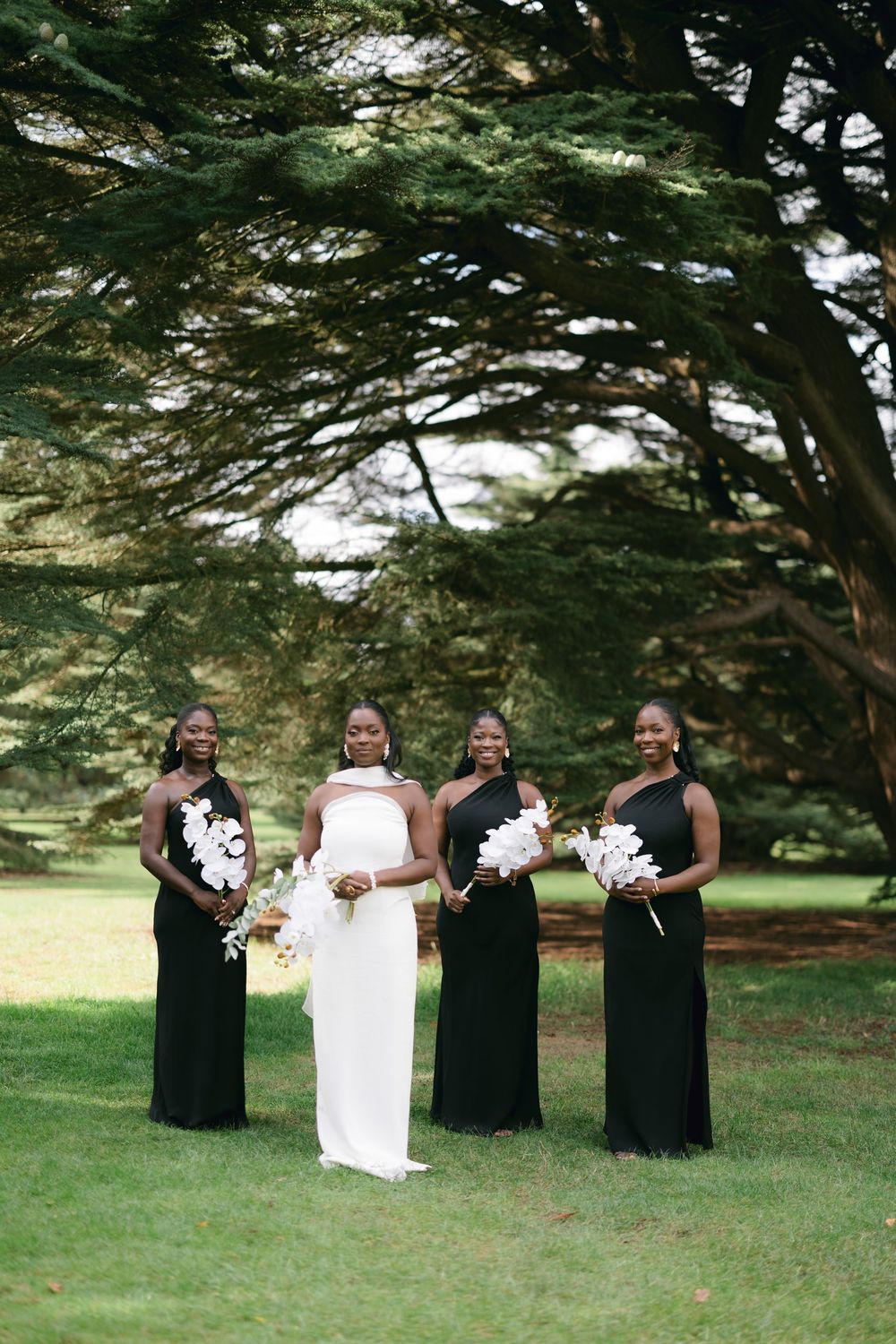 A bridal party in black dresses and white gown pose playfully together under trees in an outdoor setting.