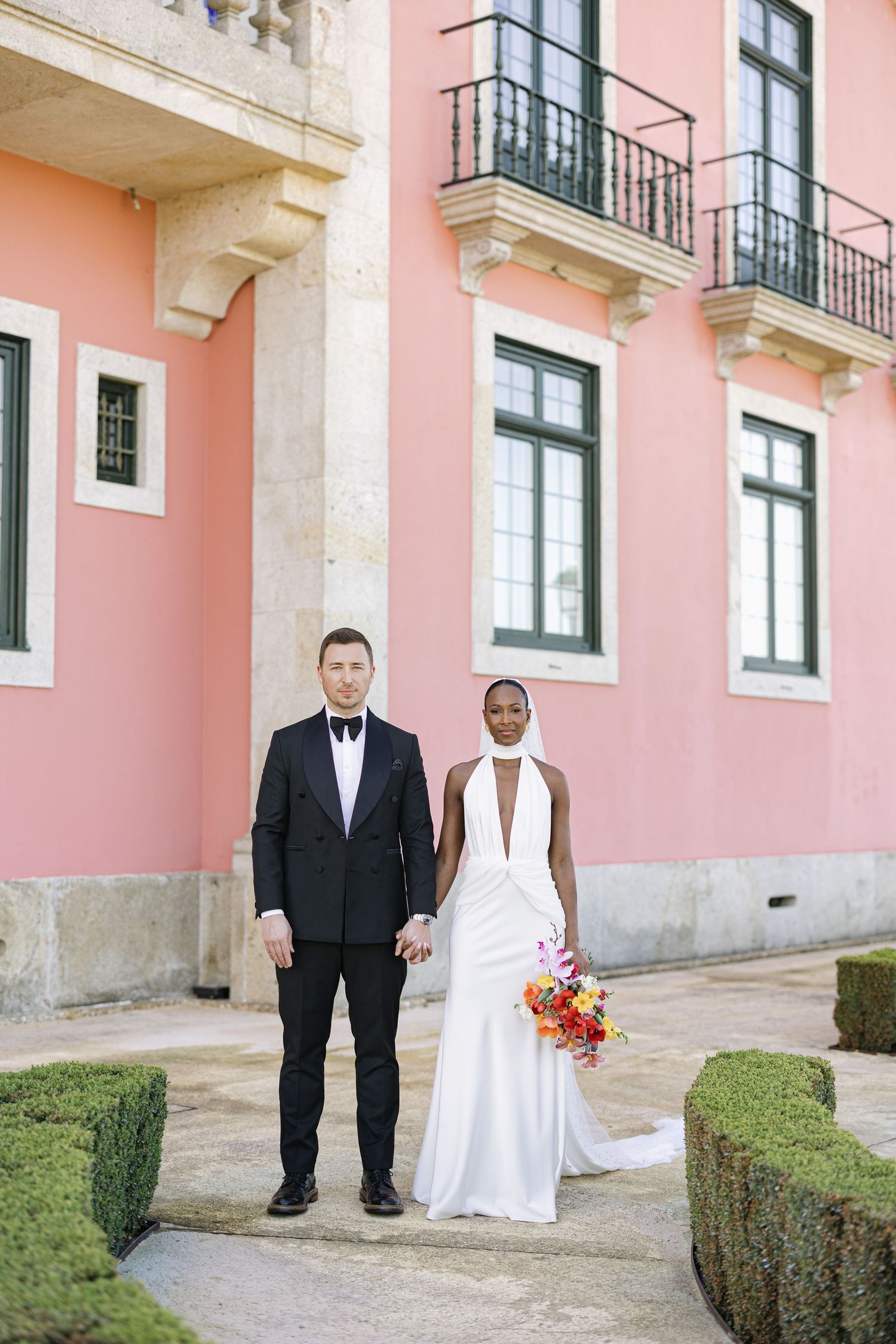 Interracial couple in wedding attire pose before a pink building with balconies in Lisbon, Portugal.