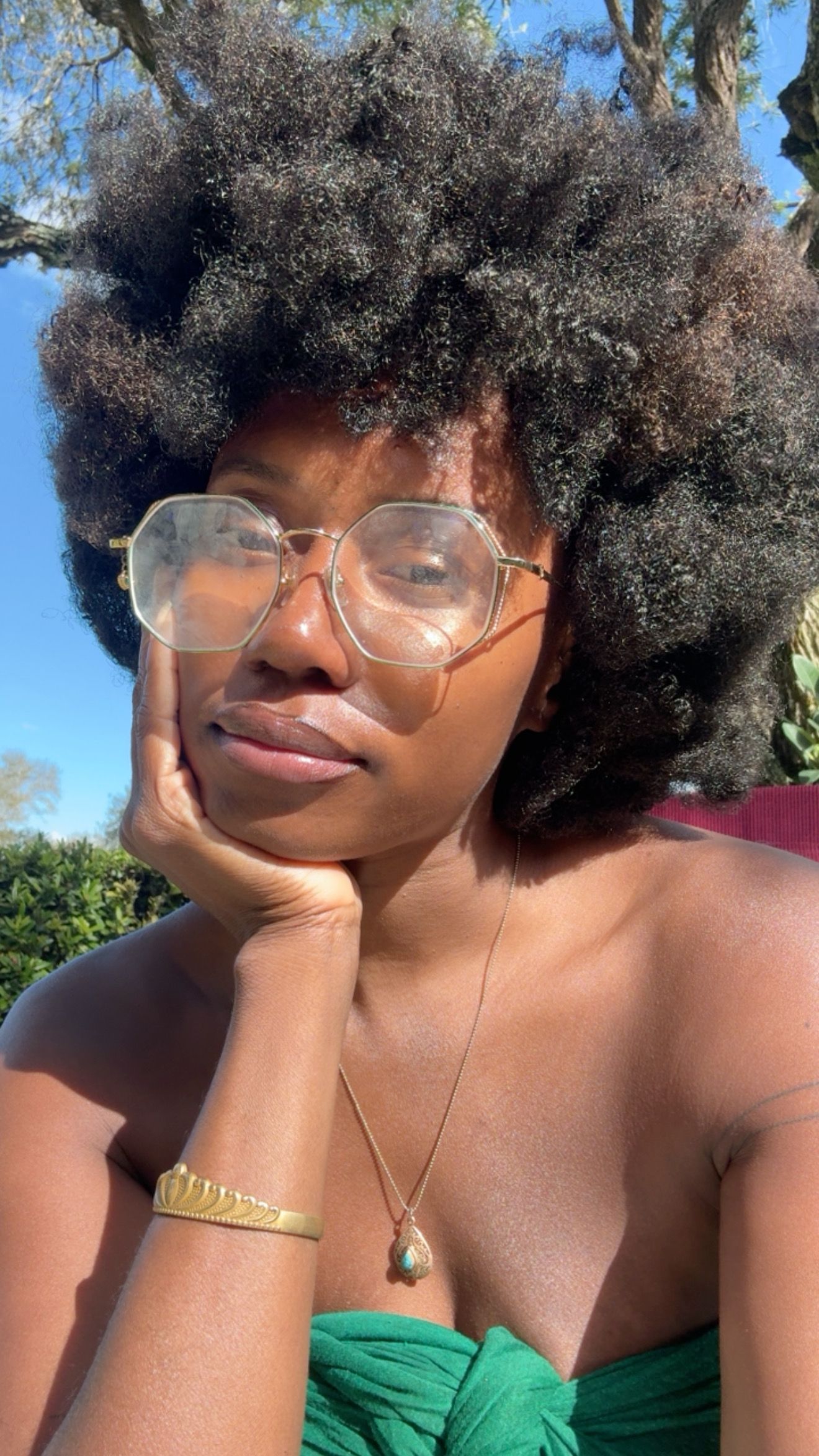 Person with natural curly hair wearing glasses and a green top poses in sunlit outdoor setting.