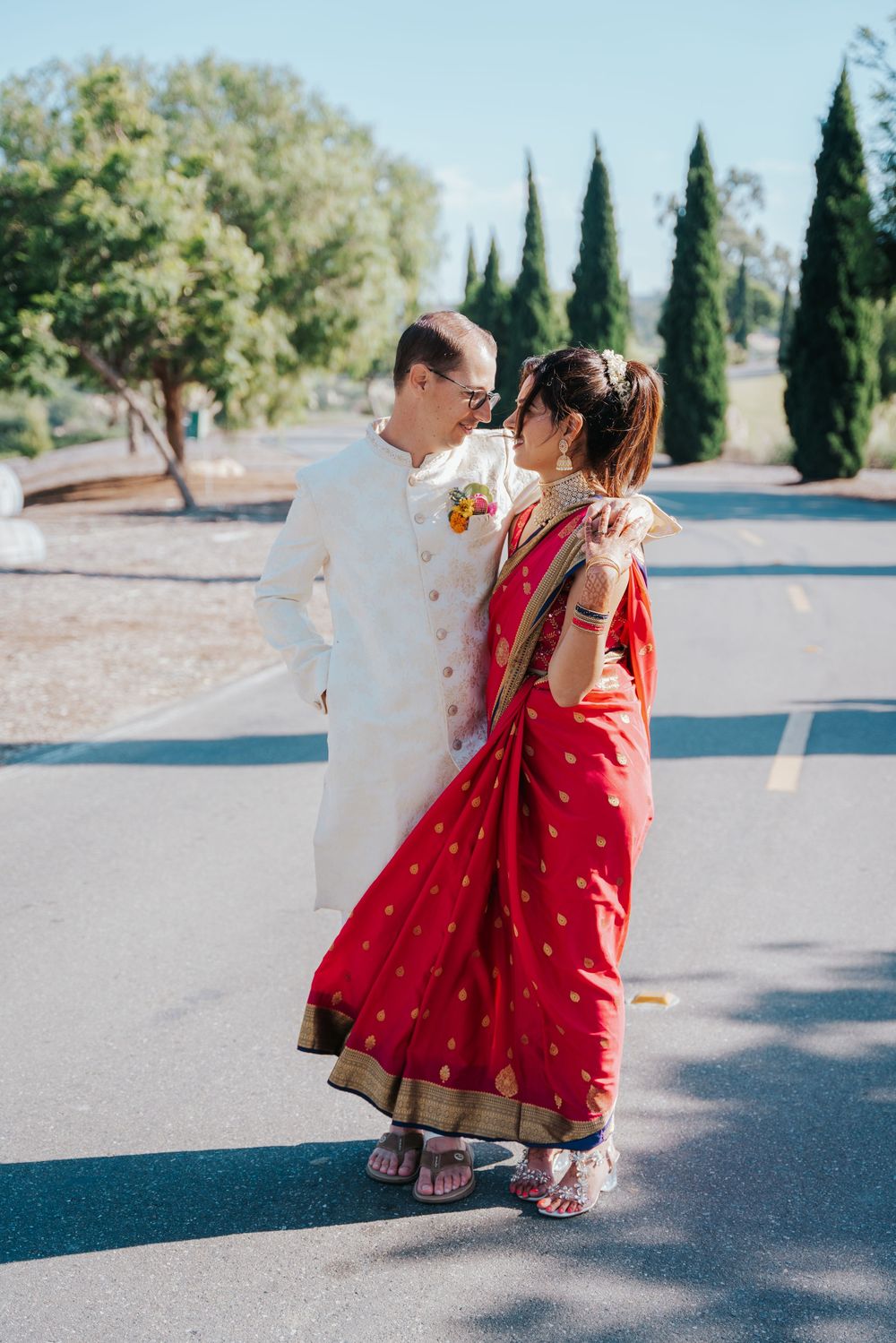 A couple in traditional Indian wedding attire, with the bride in red sari and groom in white sherwani, standing outdoors.