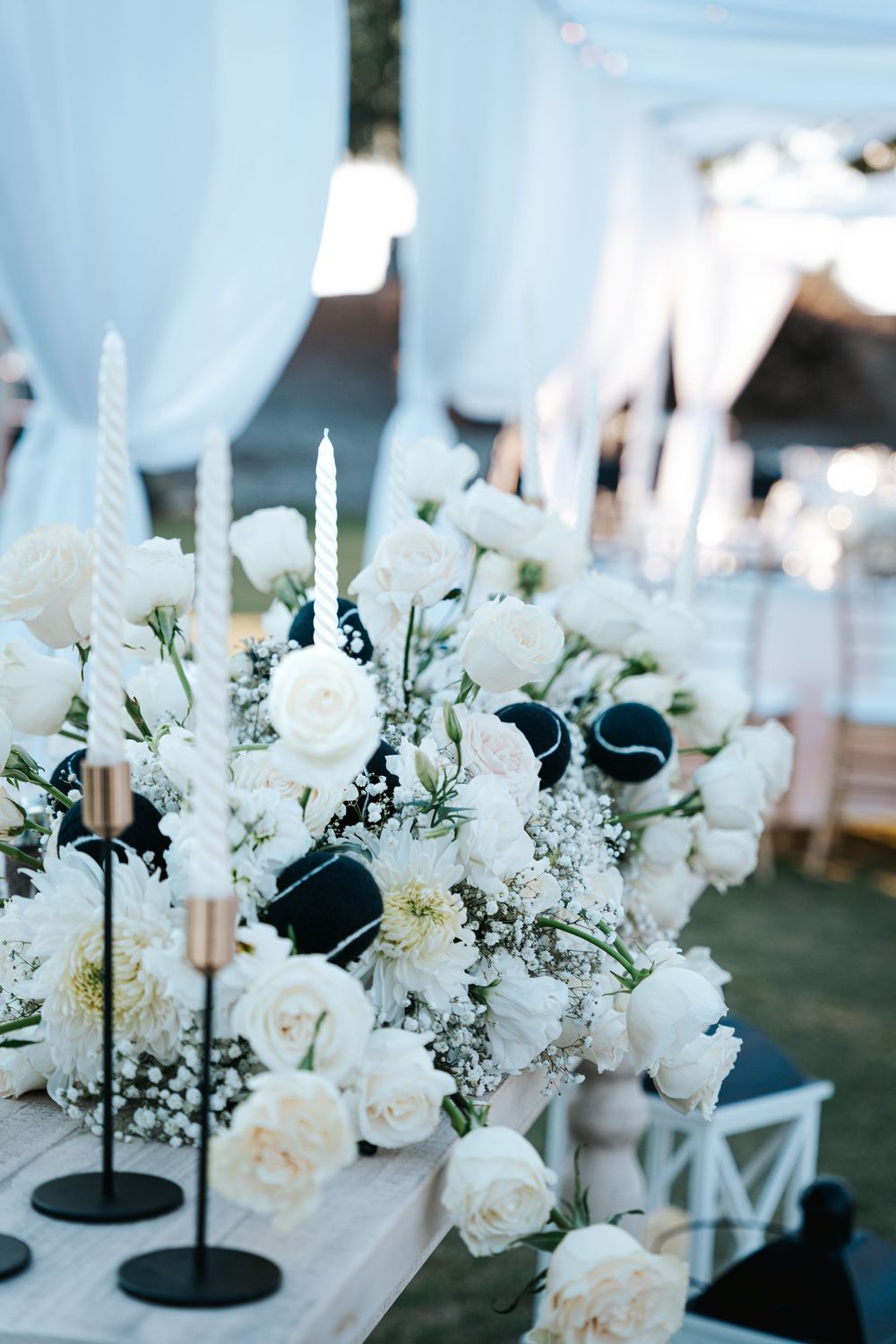 Elegant white floral centerpieces with roses and candles in black holders on a dark table at a wedding reception.
