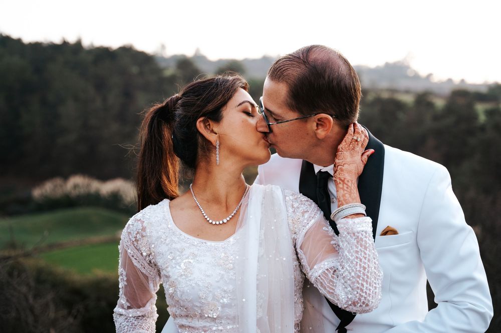 A romantic couple in wedding attire share a tender kiss outdoors with a scenic natural landscape in the background.