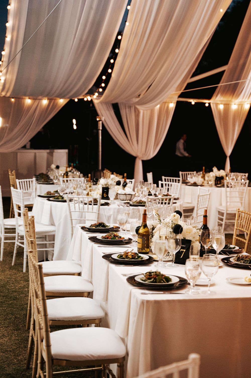 Elegant wedding reception tables set with white linens, black plates, and draped fabric ceiling with string lights.