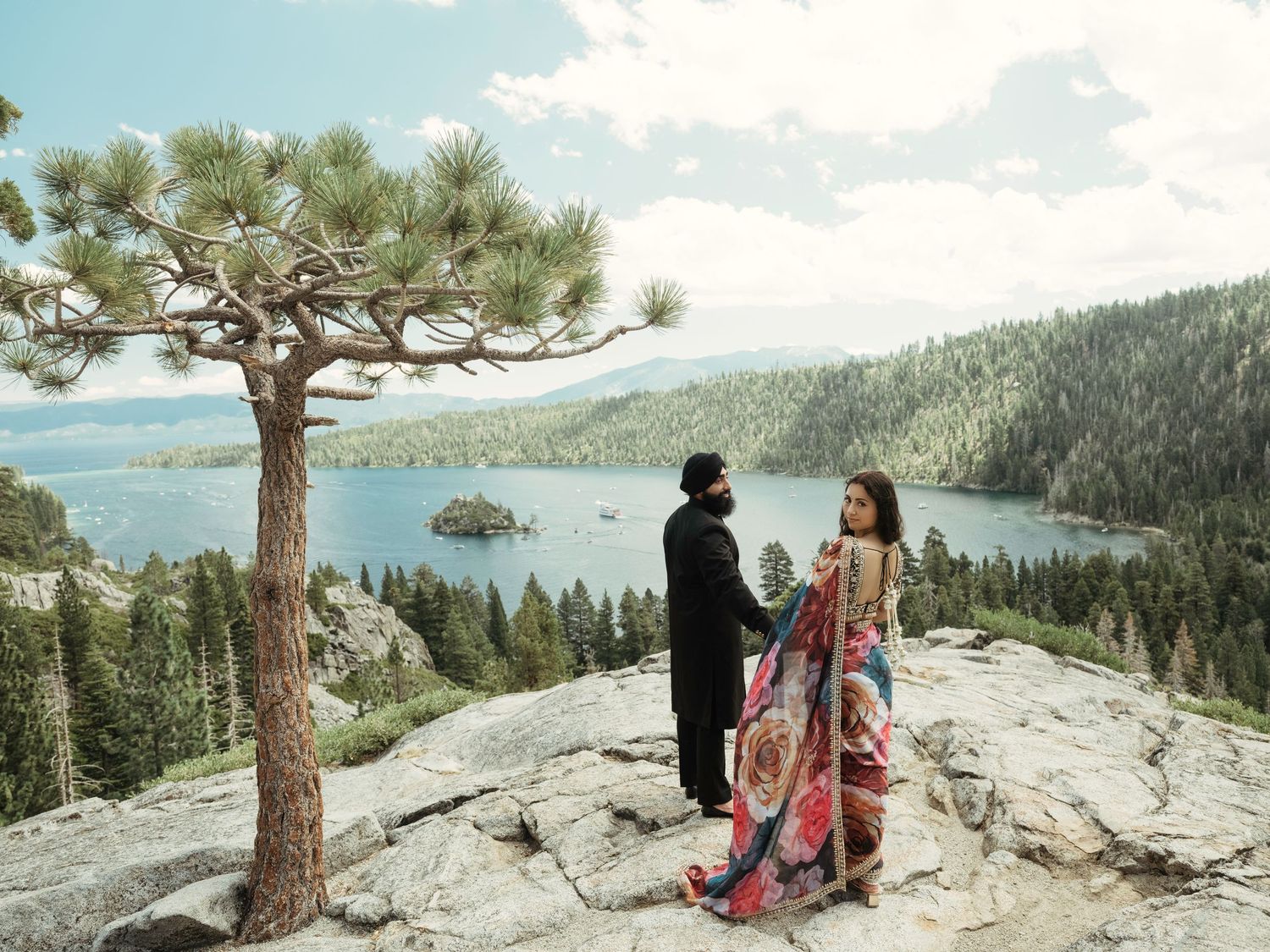 Romantic couple takes in panoramic vista of Lake Tahoe from granite cliff edge with pine trees framing the view.