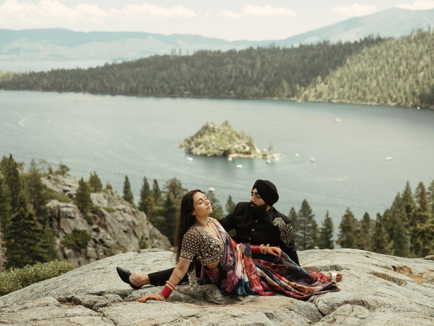 Two people relaxing on a rocky cliff edge with panoramic views of a mountain lake and forest below.