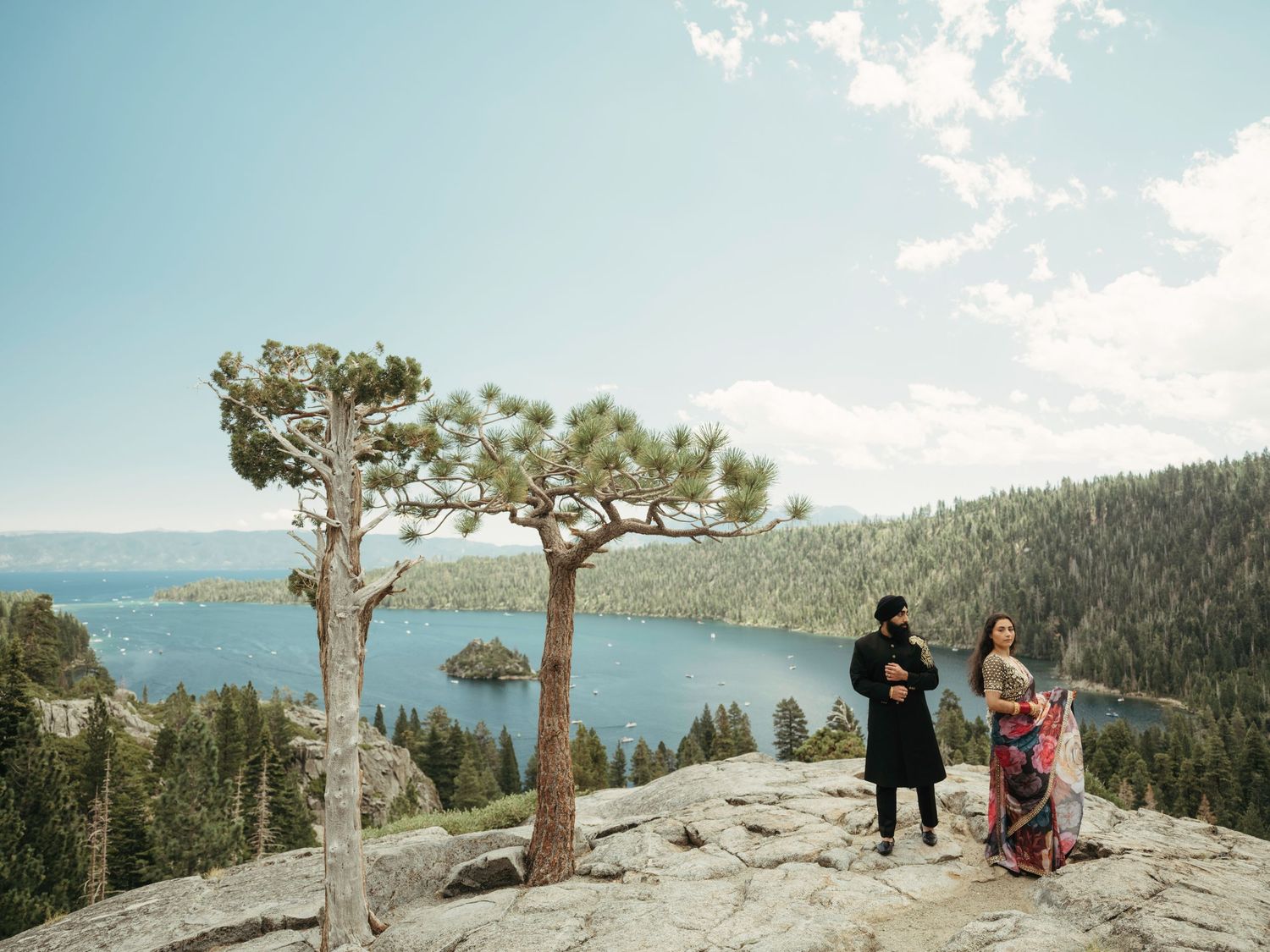 Two people stand on a rocky overlook above Emerald Bay at Lake Tahoe with tall pine trees framing the scenic view.