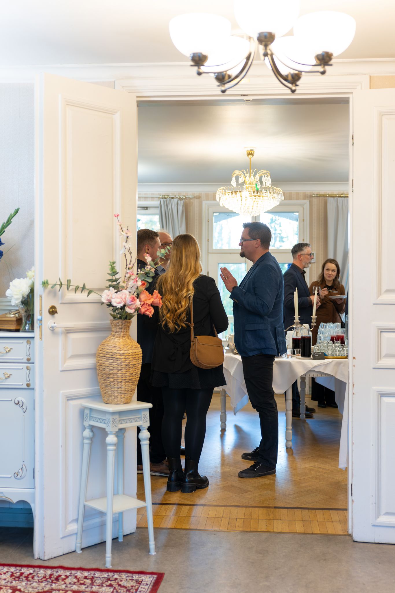 People gather in an elegant white interior space with chandeliers and hardwood floors during a social event.