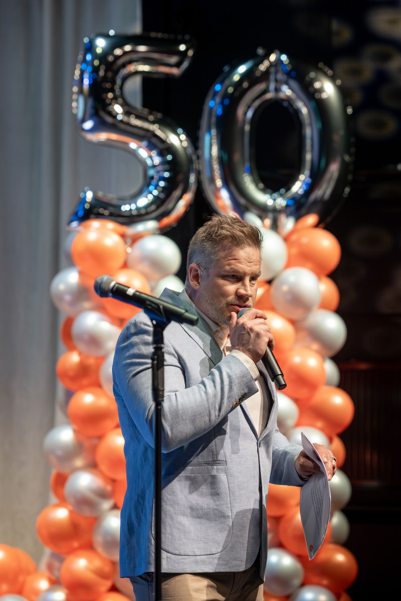 A speaker in a gray suit jacket performs at an event with decorative number 50 balloons and orange and silver balloon columns.