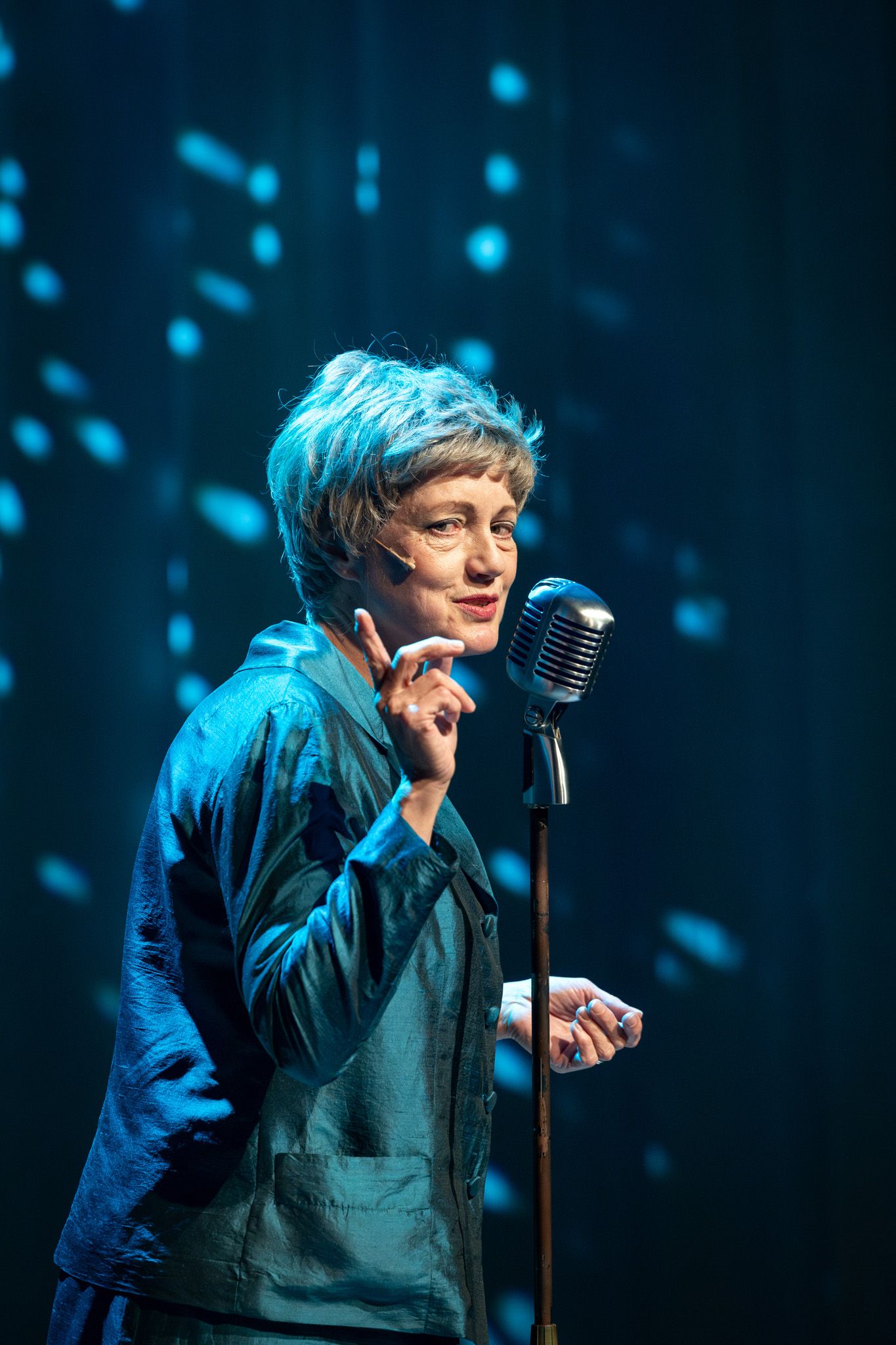 A performer in blue lighting sings passionately into a vintage microphone on stage during a concert performance.