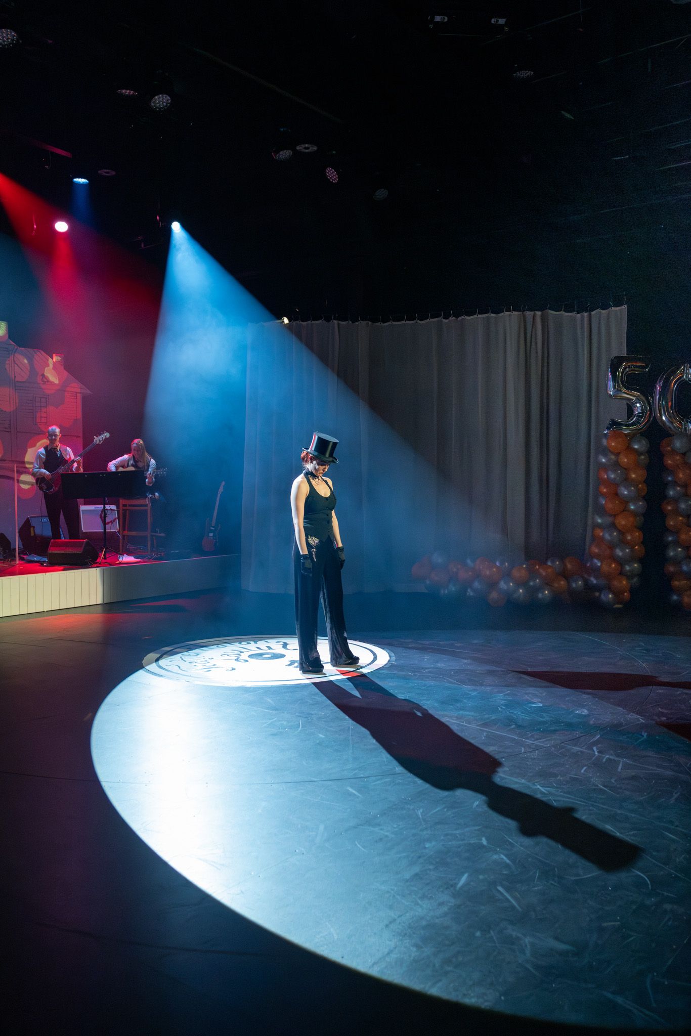 A performer stands in a dramatic spotlight on stage with blue and red theatrical lighting creating an atmospheric scene.