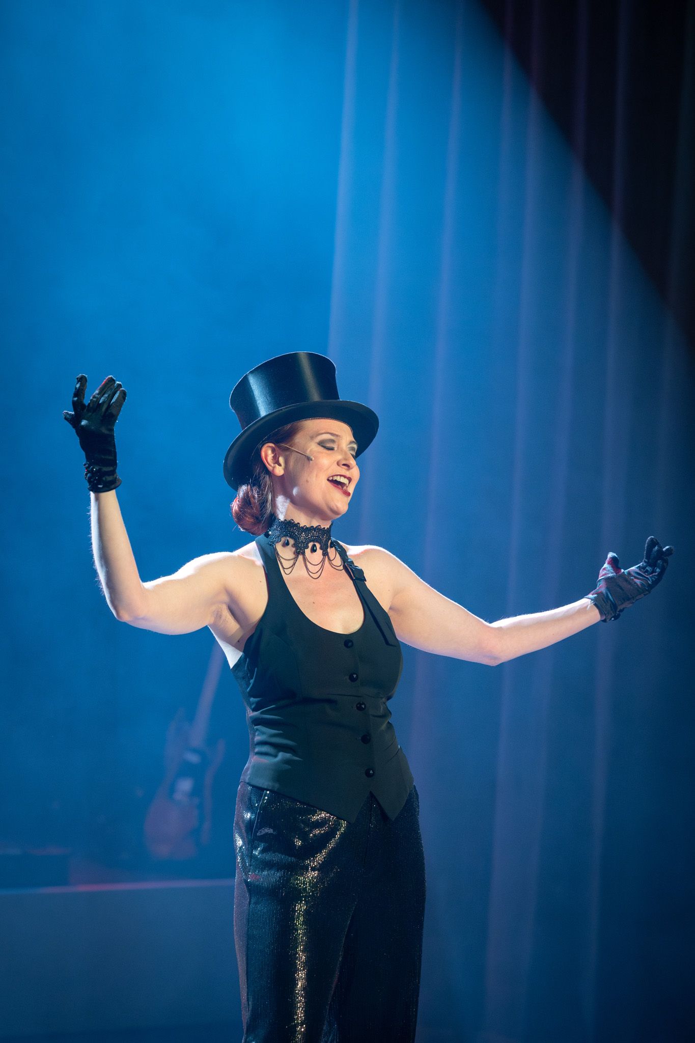 A performer in a black dress and top hat sings on stage under dramatic blue lighting during a theatrical show.