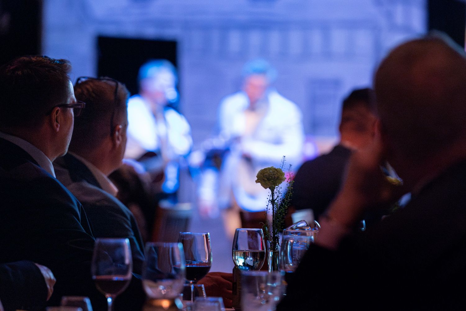 Silhouetted audience members sit in a dark room with glowing blue lighting and wine glasses on tables.