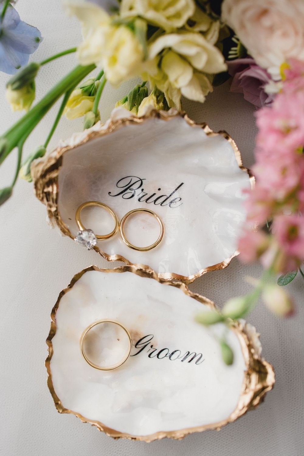 Wedding rings displayed in elegant white oyster shells with gold trim surrounded by delicate pastel flowers.