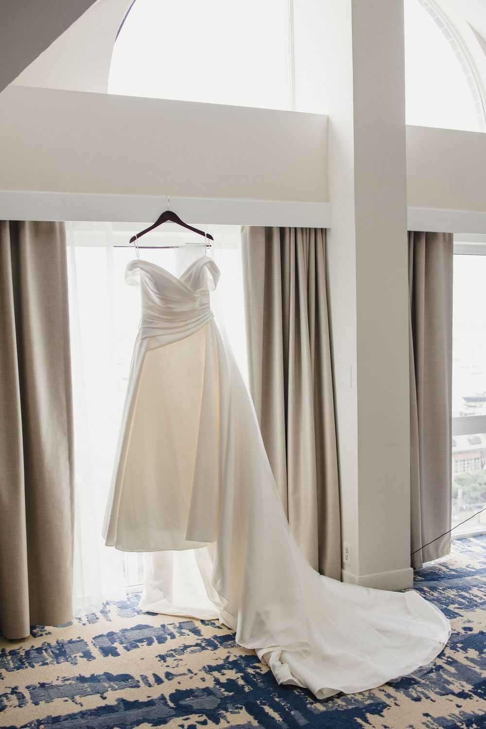Elegant white wedding dress hanging against neutral curtains in a bridal suite.