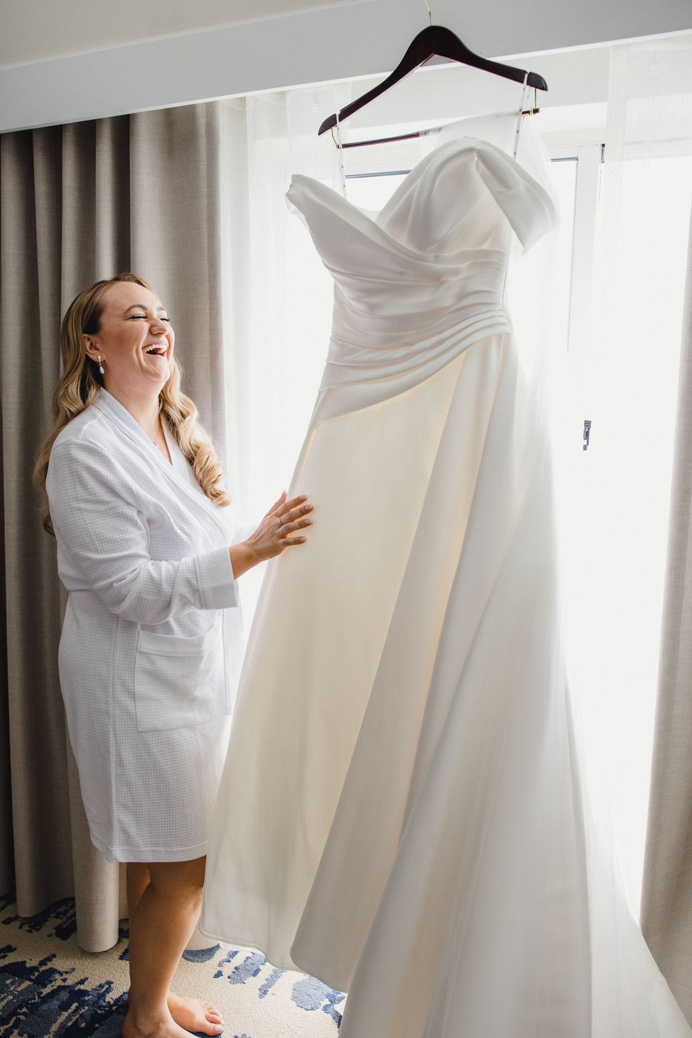 Wedding preparation scene with white bridal gown hanging by window in morning light.