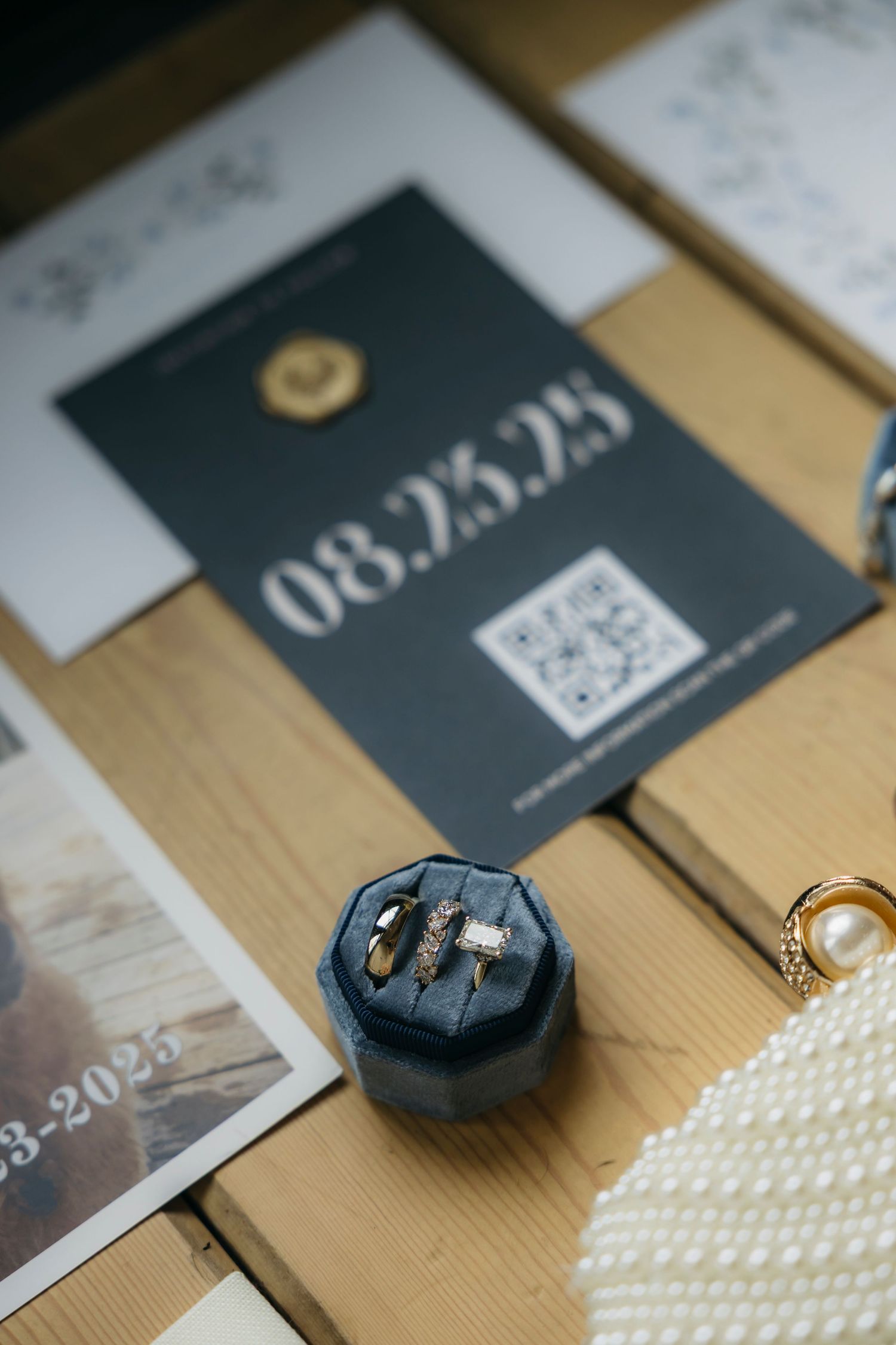 A dark velvet ring box sits next to a wedding invitation and pearl necklace on a wooden surface.