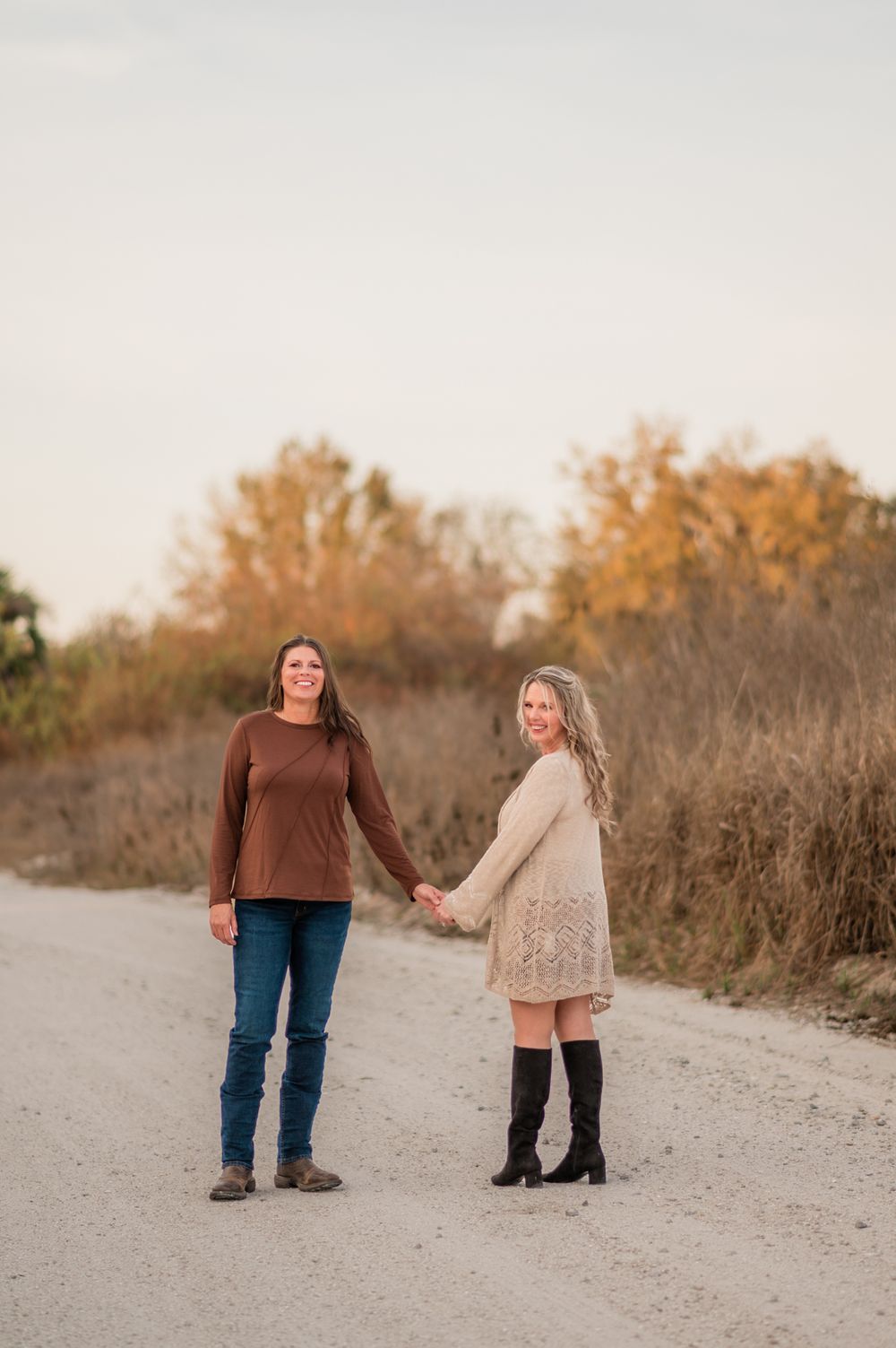 Two people holding hands while walking on a sandy path surrounded by autumn foliage at sunset.