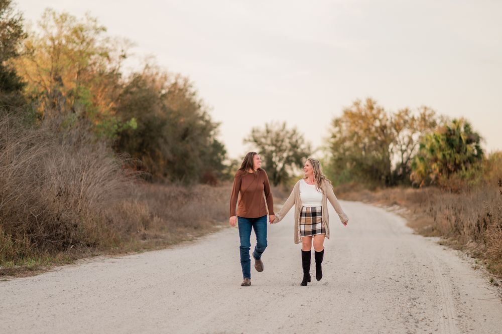 Two people walk hand in hand down a gravel road surrounded by autumn trees at sunset.