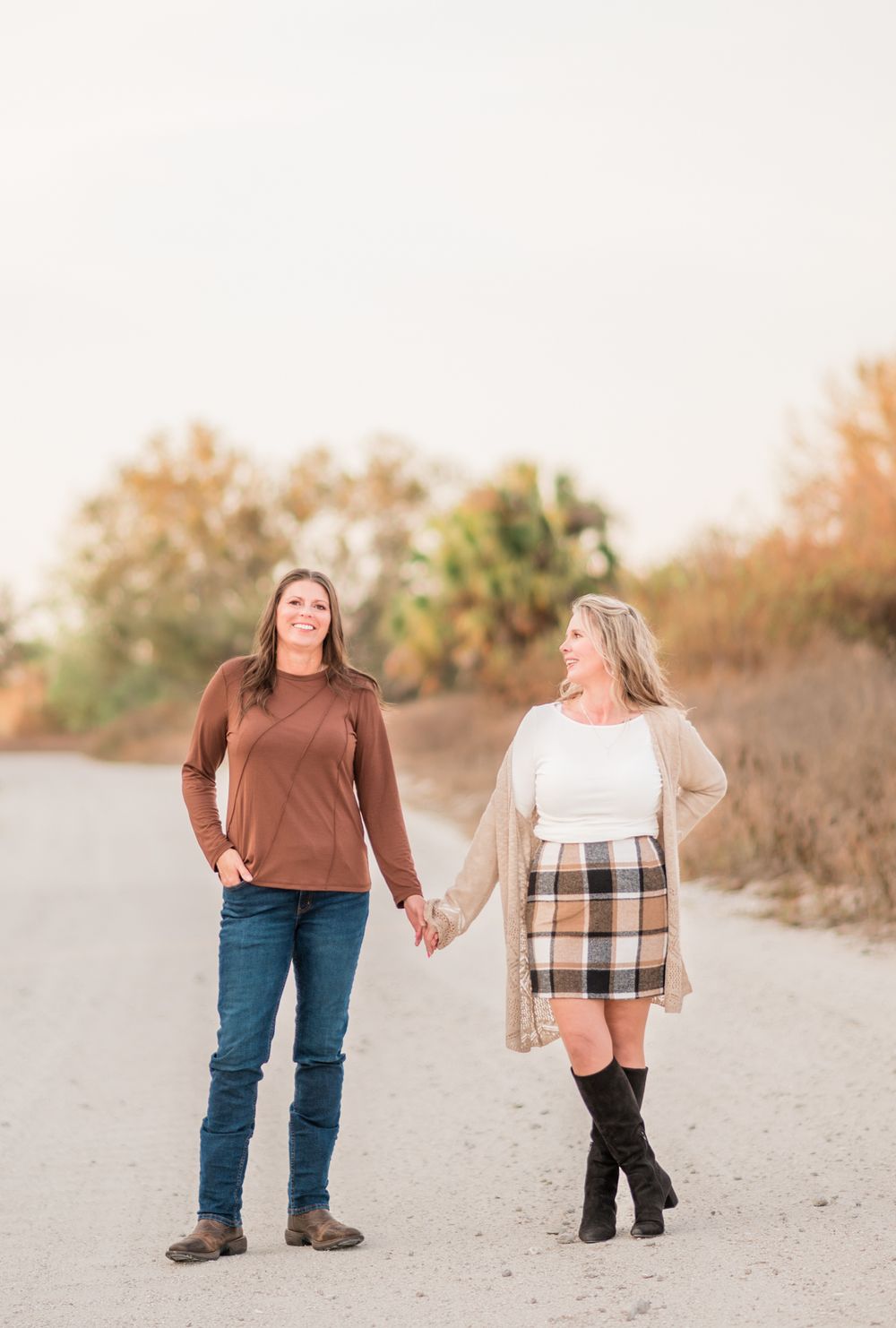 A couple walking hand in hand on a beach path during golden hour with autumn foliage in the background.