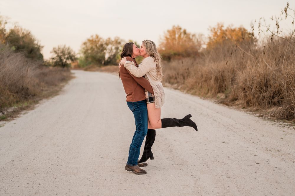 Two people playfully interact on a sunlit trail surrounded by dry brush and fall colors.